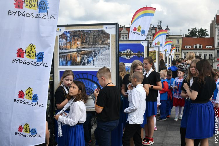 Dzieci oglądają plenerową wystawę zdjęć. Na proporcach logo Bydgoszczy z zabytkowymi spichrzami.
