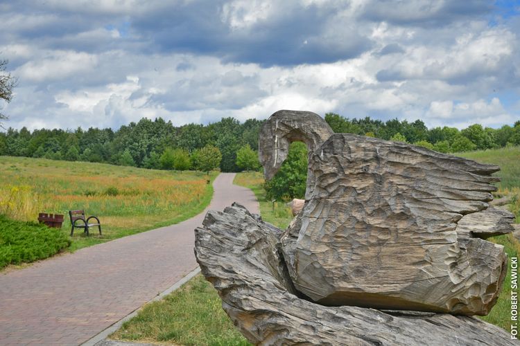 Wyłożona kostką ścieżka w parku miejskim. Z boku drewniana rzeźba przedstawiająca łabędzia.